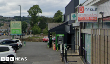 A strip of shops on a main road. In the foreground is a For Sale sign on an empty building. Behind is a pharmacy and an Asda.
