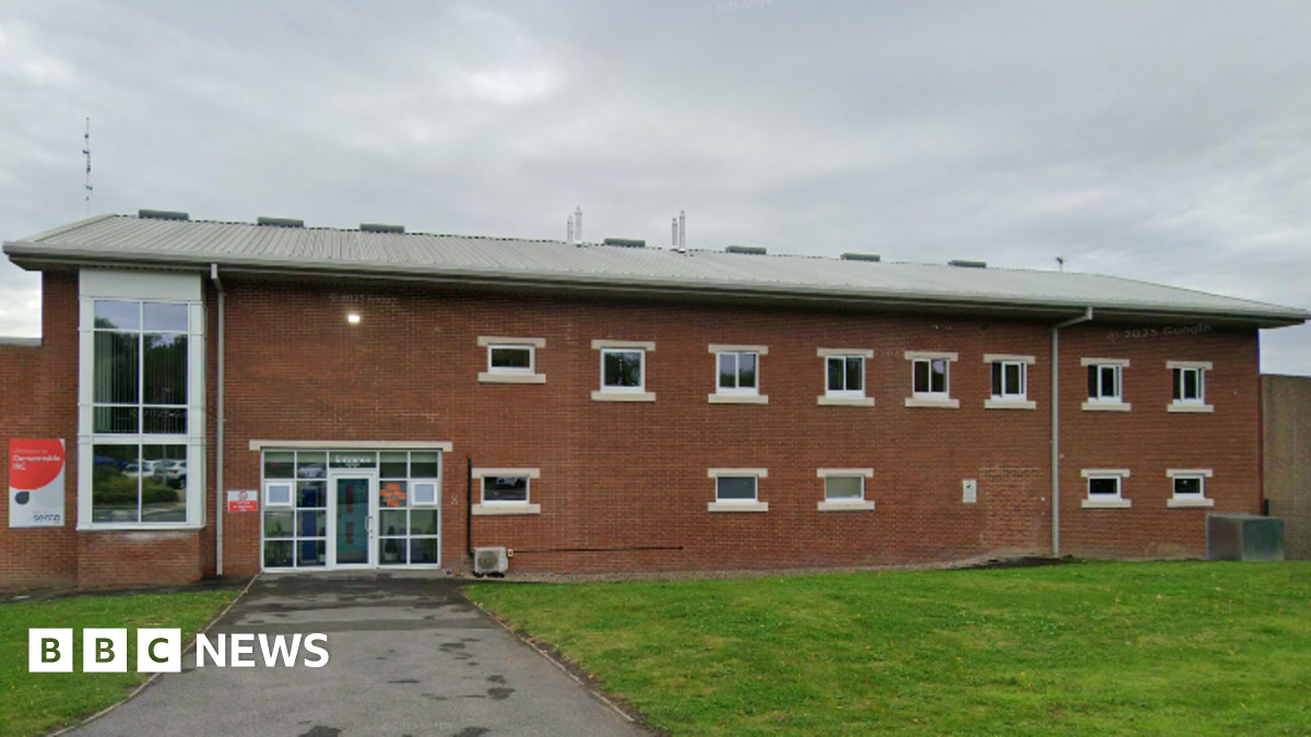 Derwentside Immigration Removal Centre is a two-storey red brick building with small, white-framed windows on both floors. To the left is a much larger long window spanning both floors next to the glazed entrance that has a single central door. The walls of the complex stretch out either side and the roof of another building stands behind the wall. The base of a flagpole can be seen in the foreground.