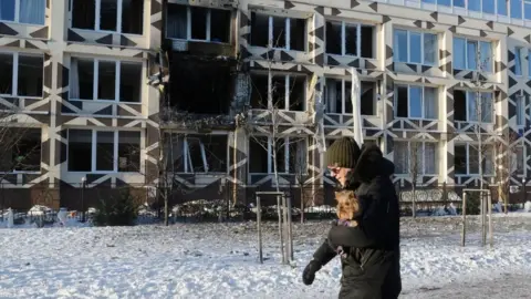 EPA A man holding a dog walks near the site of a Russian drone strike on a private medical clinic. He has a hat and gloves and is walking on snow.