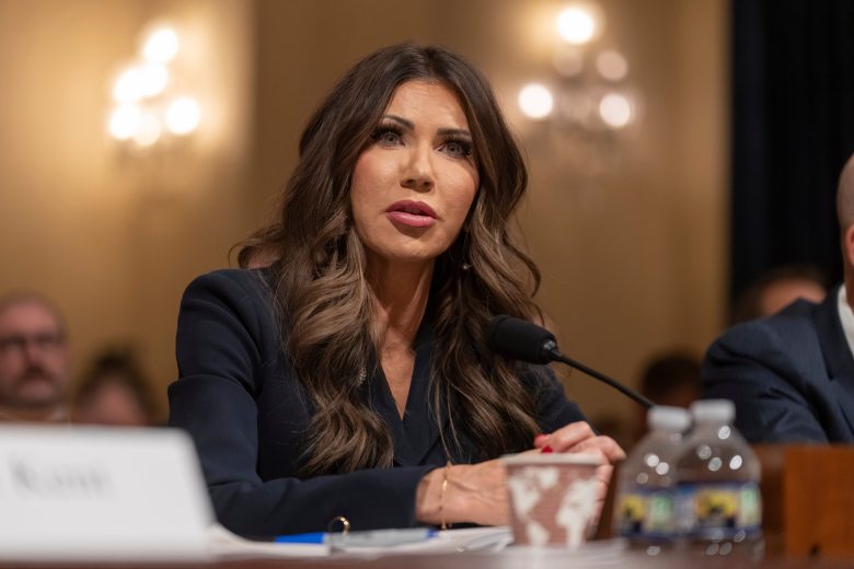 A person sitting at a table with folded hands, speaking into a microphone