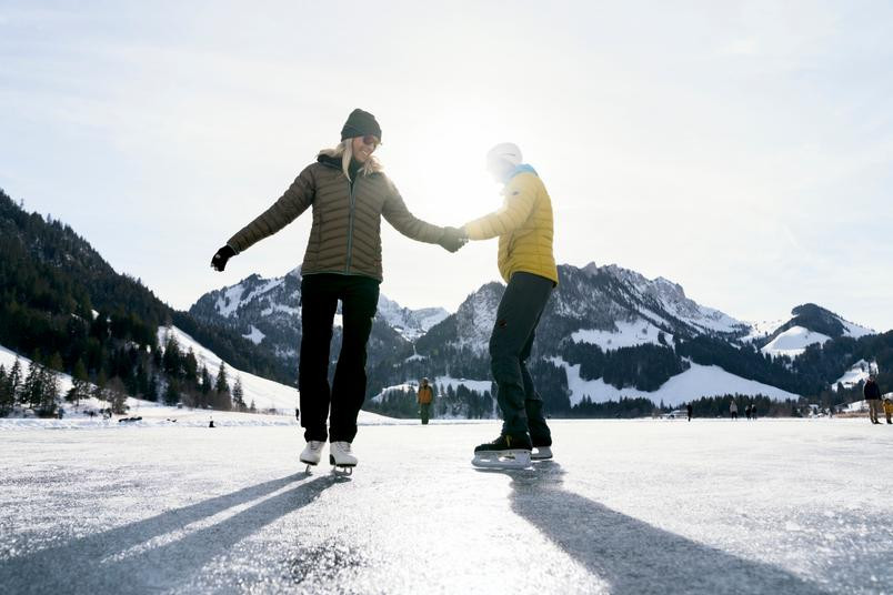 Deux personnes patinent main dans la main sur un lac gelé entouré de montagnes enneigées.