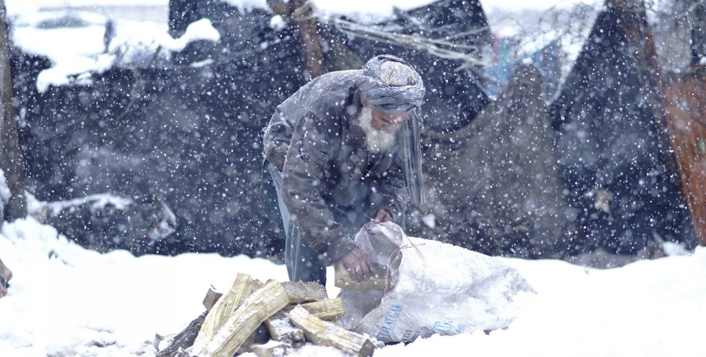 An elderly Afghan man struggles through heavy snowfall while trying to start a fire for warmth, highlighting the harsh winter conditions faced by millions in need of humanitarian assistance across Afghanistan. (Photo Credit: OCHA Afghanistan)