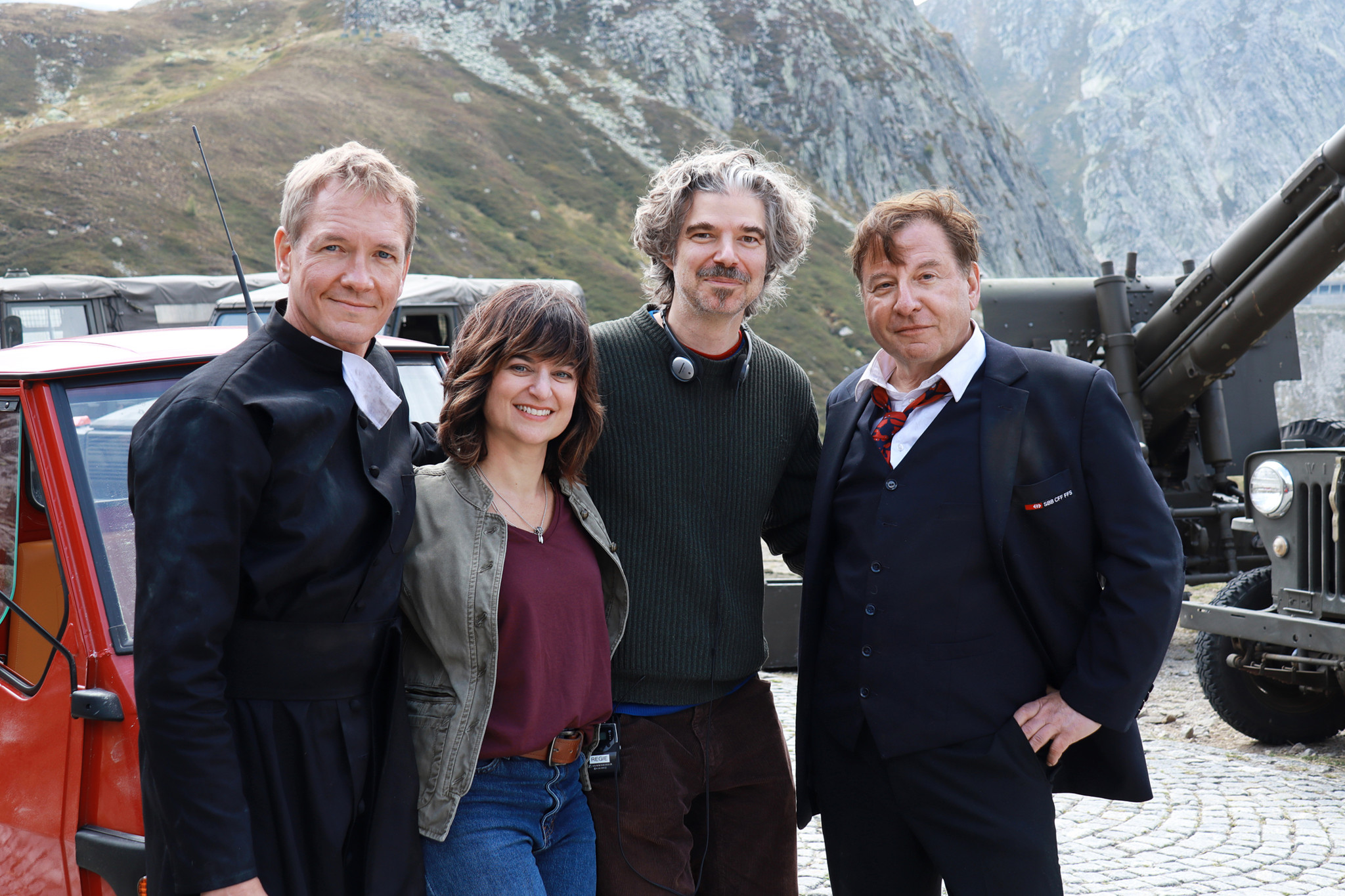 Vincent Kucholl, Catherine Papagni, Regisseur Peter Luisi und Beat Schlatter posieren vor einer malerischen Berglandschaft im Ticino. Im Hintergrund ist ein rotes Auto und ein antikes Geschütz zu sehen.