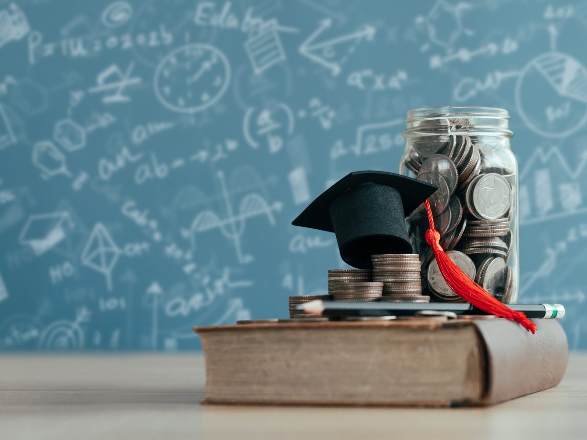 An old book with a college cap, coins and a money jar atop it, in front of a chalk-covered board.