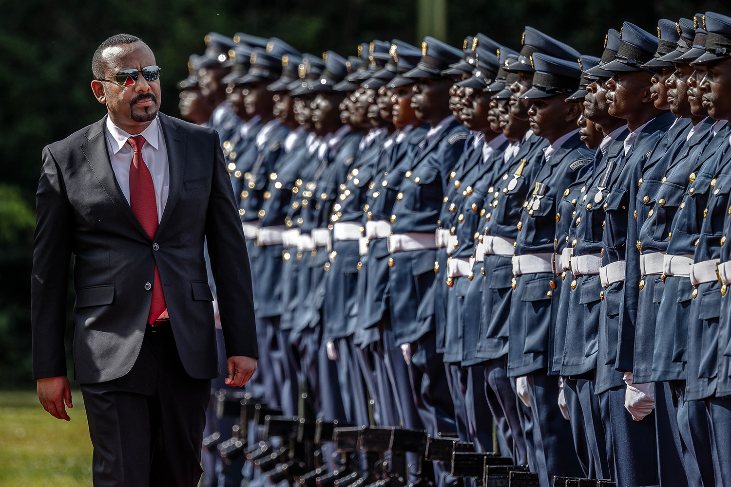 Prime Minister of Ethiopia Abiy Ahmed, wearing a suit and sunglasses, walks past uniformed military personnel standing at attention in formal dress uniforms.