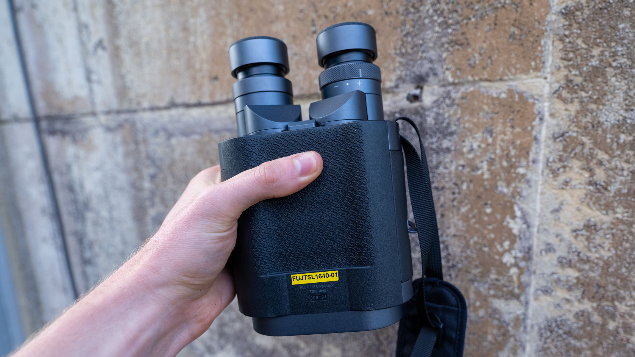 A hand holding the Fujifilm Techno-Stabi TS-L 1640 image-stabilized binoculars with a beige wall in the background.
