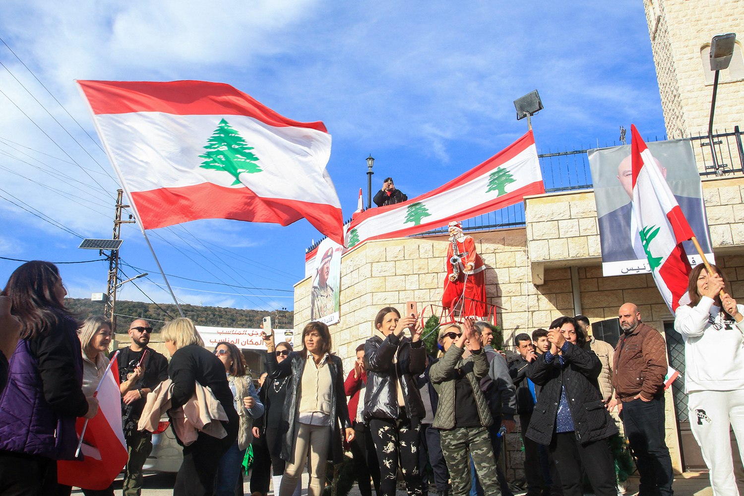 A crowd gathers in front of stone buildings under a bright blue sky, with political posters and visible on the buildings. People wave Lebanese flags.