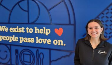 a woman wearing a black shirt that says Butterball stands in front of a royal blue wall with wording on it that says "We exist to help people pass love on."