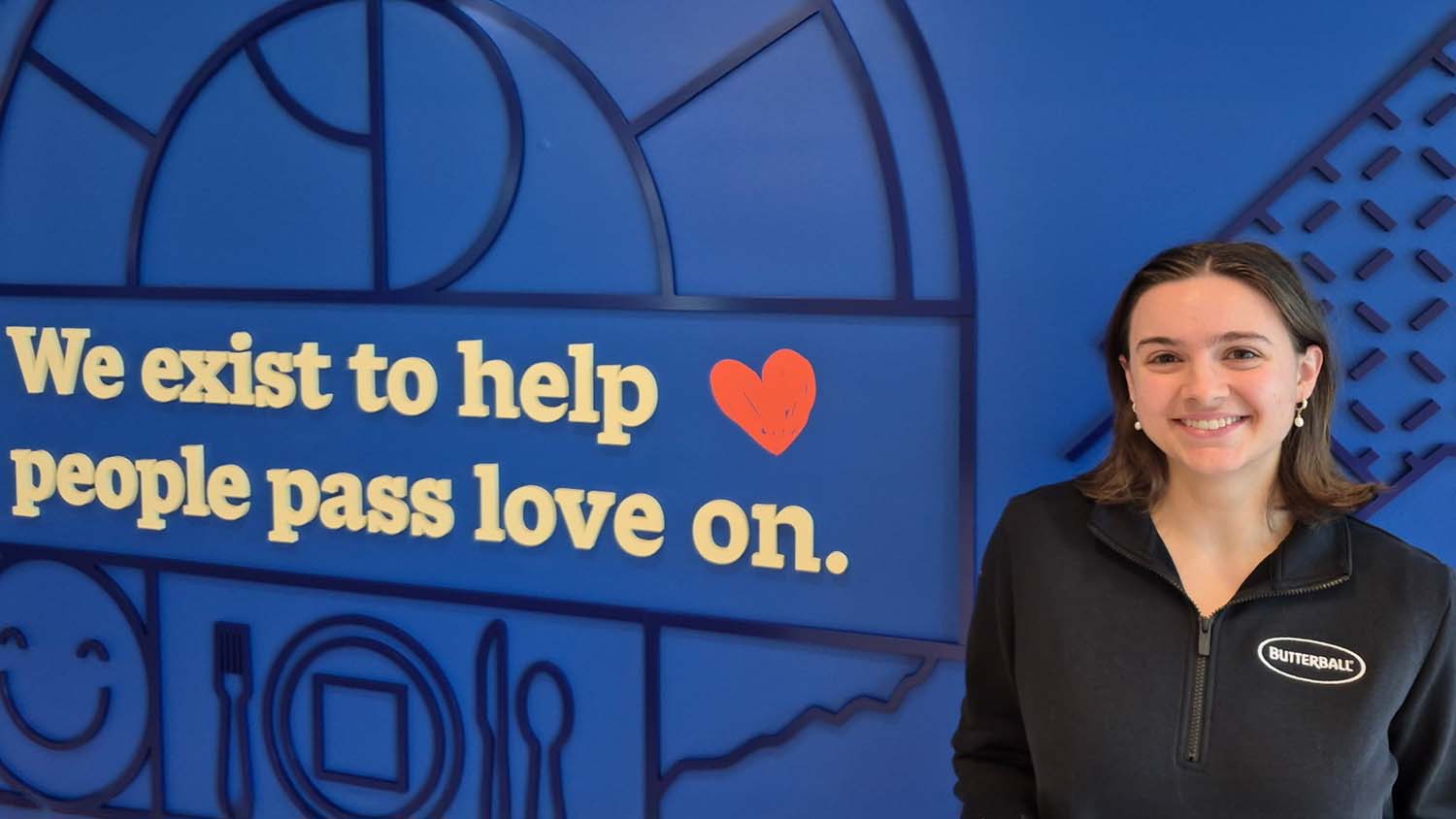 a woman wearing a black shirt that says Butterball stands in front of a royal blue wall with wording on it that says "We exist to help people pass love on."