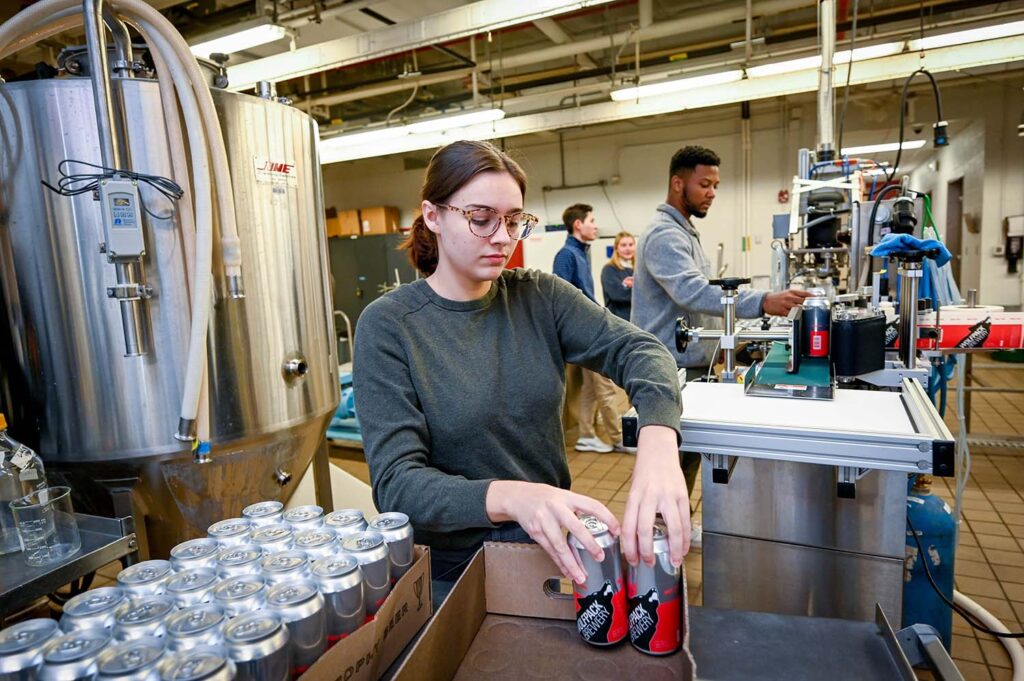 a woman in a brewing lab puts cans that say "Wolfpack Brewing" in a box