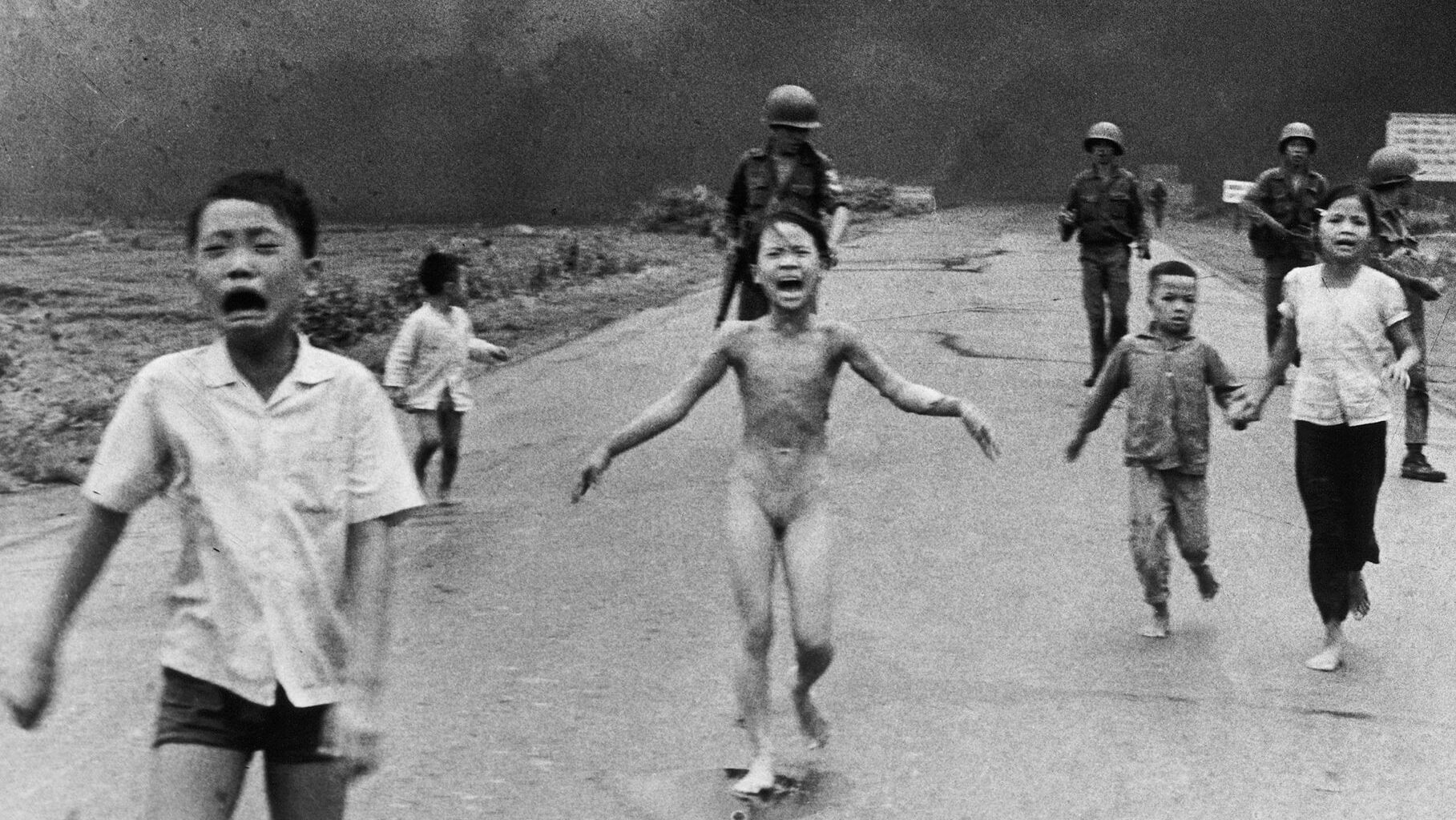 A group of children, some crying and barefoot, run down a road toward the camera with smoke and armed soldiers in the background. The central child appears distressed and unclothed.