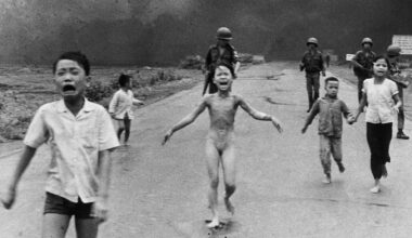 A group of children, some crying and barefoot, run down a road toward the camera with smoke and armed soldiers in the background. The central child appears distressed and unclothed.