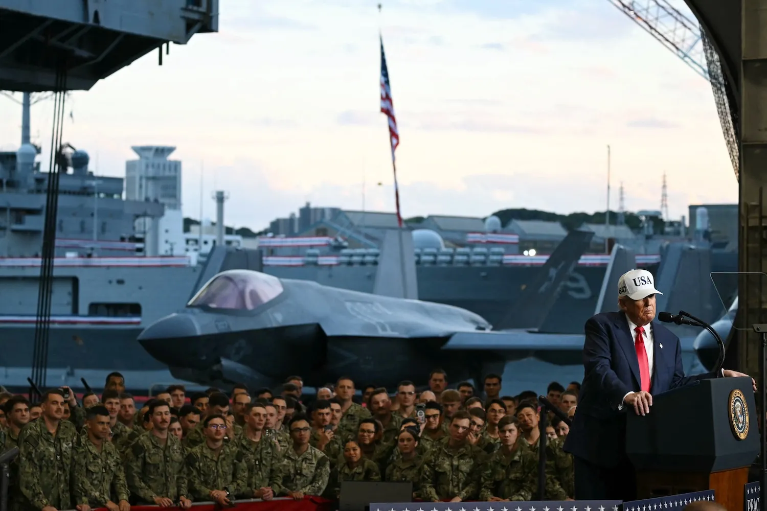 Trump stands at a podium wearing a white baseball cap that reads "USA." Behind Trump, rows of military personnel in uniform stand and watch him. A military jet sits behind the crowd.