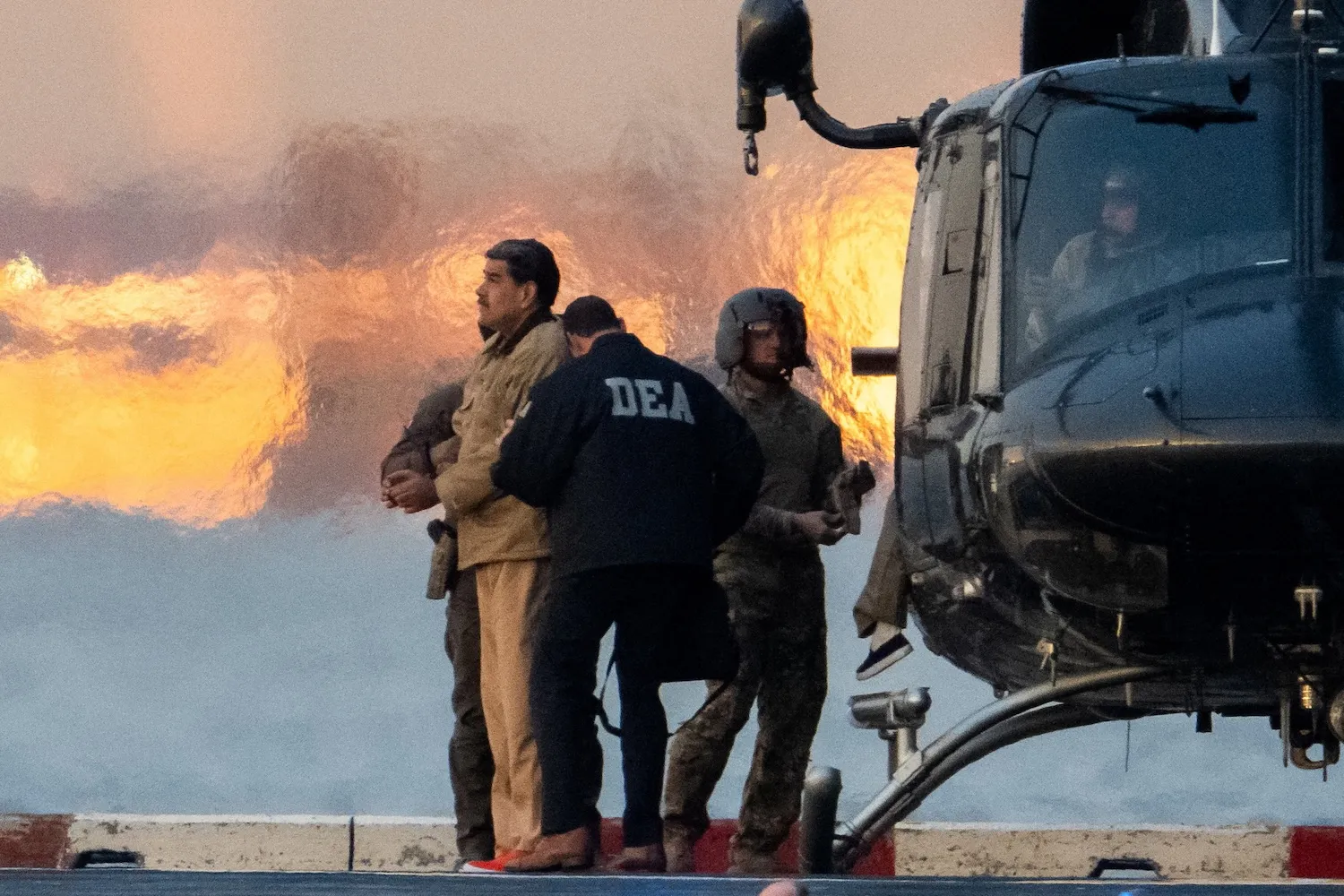 Venezuelan President Nicolás Maduro stands and gazes ahead while being held by military personnel and a man wearing a jacket that reads "DEA." They stand beside a helicopter.