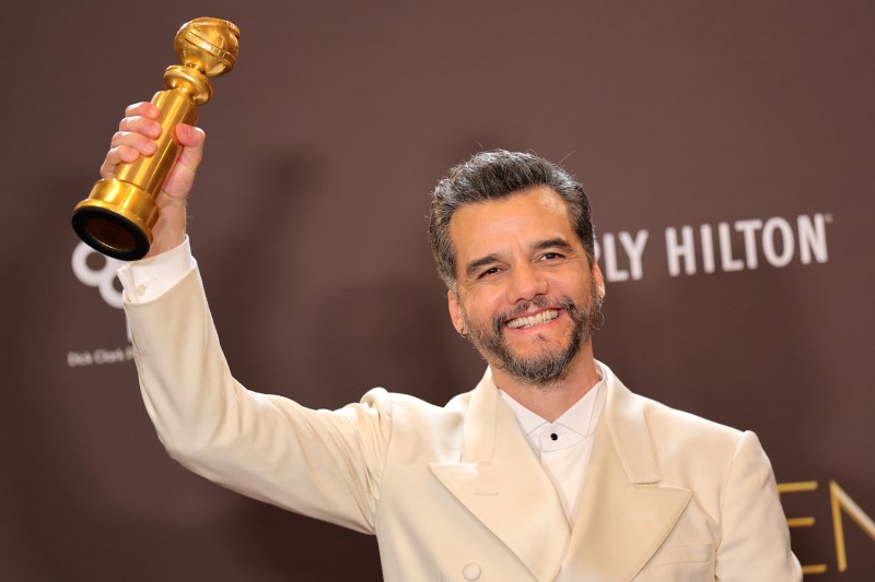 Against a brown backdrop, actor Wagner Moura holds a Golden Globe award in his right hand. He is wearing a cream-colored suit jacket.
