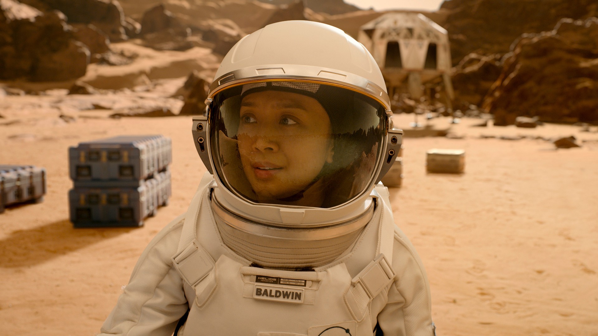 Close up of a female astronaut on Mars. In the background there are several containers and a white building. This is a screenshot from the Apple TV series "For All Mankind"
