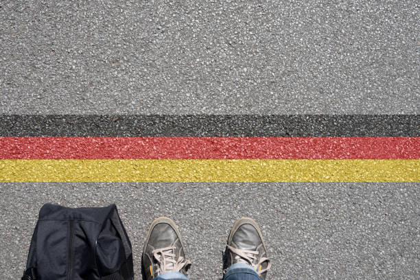 Person stands at a road marking coloured like the German flag, seen from above with shoes and a bag at the border line.