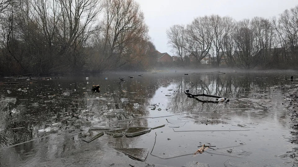A pond with frozen shards of ice floating on it. The sky is grey and there is haze on the horizon, where homes can be seen in the distance. The pond is surrounded by trees.