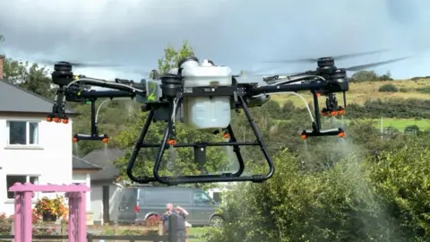 A spraying drone lifting off from a garden and spraying water. The drone is black on the four arms and under carriage. In the middle is a silver box. 