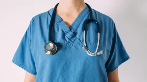 Getty Images A person is standing facing the camera wearing blue hospital scrubs with a stethoscope around their neck. You cannot see their face or any defining features. 