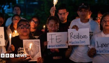 Millie Bobby Brown and David Harbour in character as Eleven and Jim Hopper. Eleven wears red shorts over grey leggings and a grey jumper. Jim Hopper wears a black leather jacket, a black beanie hat and dark trousers.They are speaking to director Frank Darabont on the set of Stranger Things. There is a vintage advertisement posted in the background with debrief on the ground.