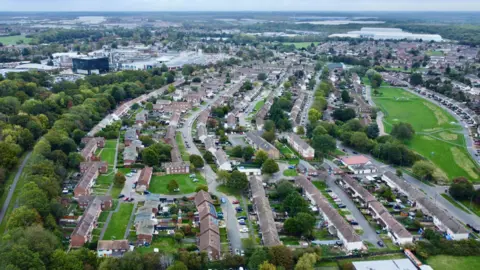 Ant Saddington/BBC A drone image of a housing estate, showing rooftops, gardens, surrounding green space and roads. In the distance are industrial units. 
