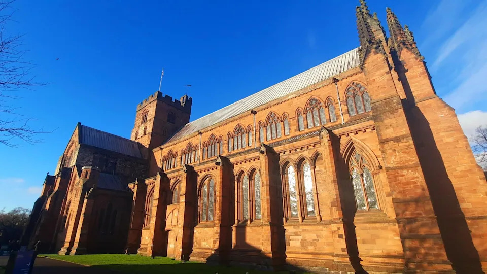 A general view of Carlisle Cathedral taken from its side. The building in red sandstone has arched windows all along its side, with gothic-type detailing and turrets. The sky is blue and bright.