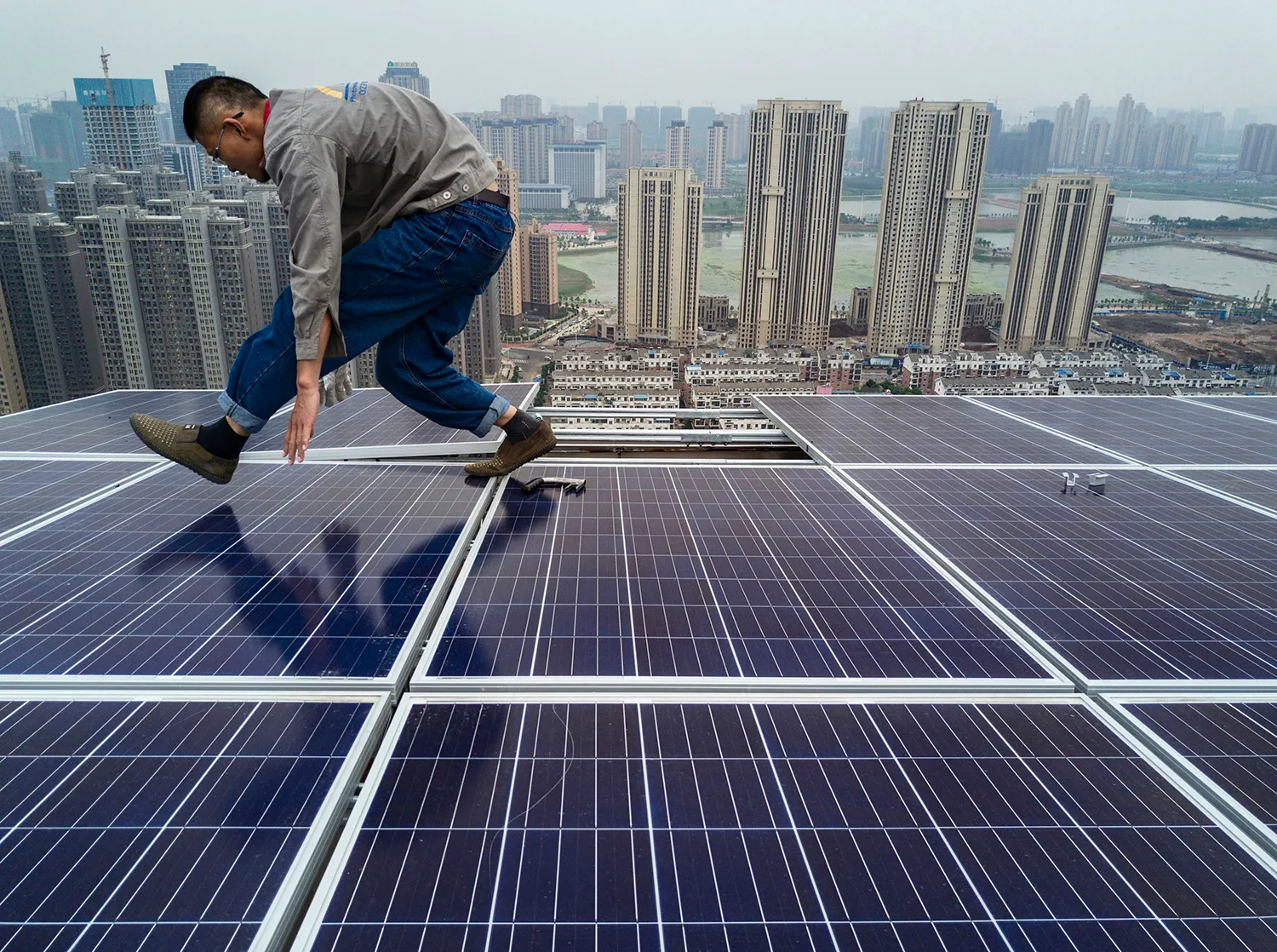 A man bends over as he walks atop solar panels atop a tall building with other skyscrapers in the background.