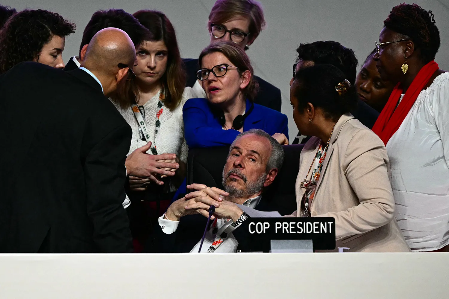 A man seated with his hands folded behind a "COP President" sign with multiple people clustered behind him talking.
