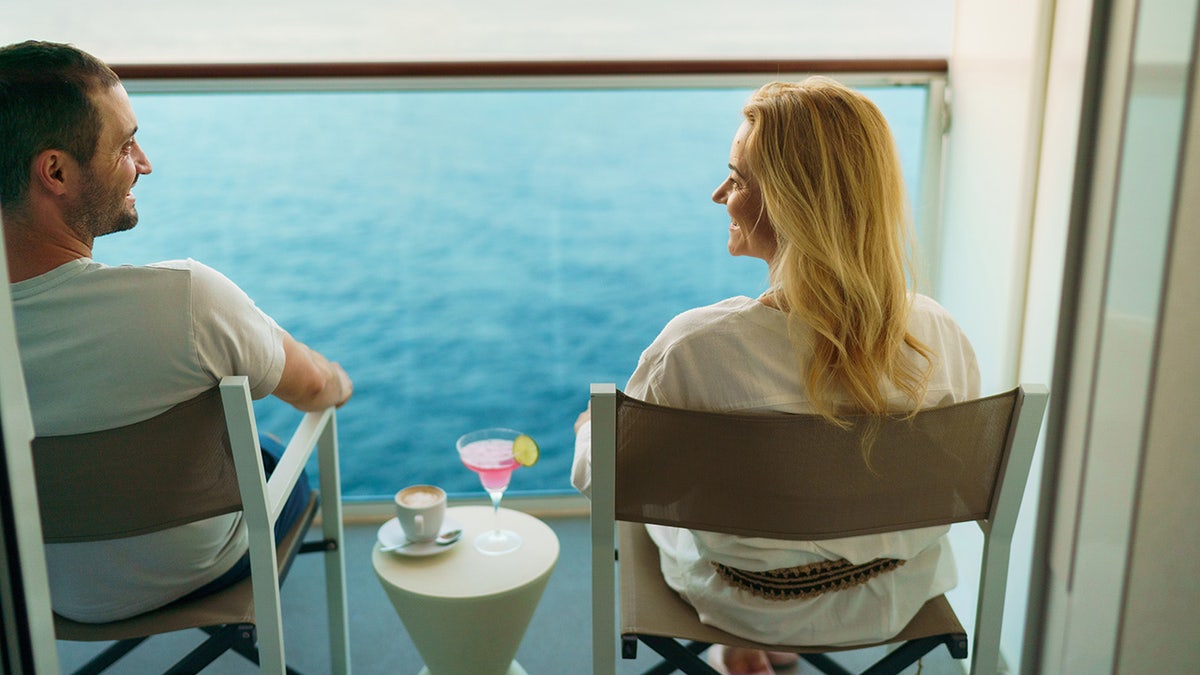 A couple sitting on deckchairs on the balcony of the cruise ship smiling and enjoying their drinks with seas in background.
