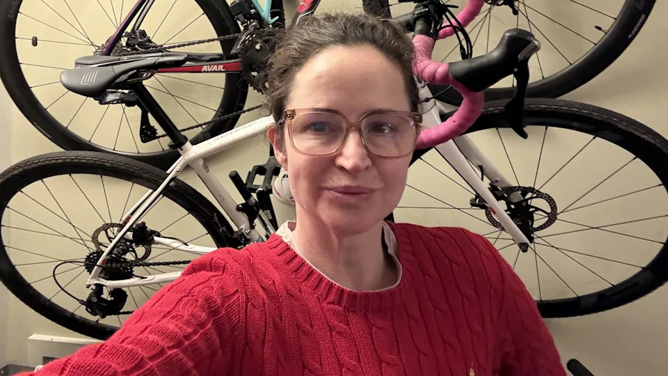 Wearing a red jumper and glasses, Kate Corden, stands in front of bicycles hanging on a wall.