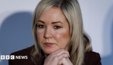 A close-up of Michelle O'Neill during a press conference at the British-Irish Council in Wales in December 2025.  She is a woman with straight, shoulder-length blonde hair.  She is wearing a dark suit jacket and is resting her face on her hands, as though listening to someone speak.