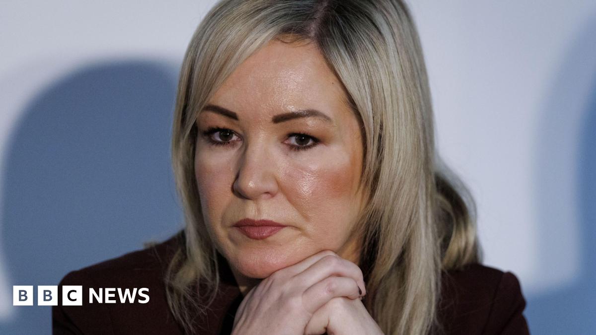 A close-up of Michelle O'Neill during a press conference at the British-Irish Council in Wales in December 2025.  She is a woman with straight, shoulder-length blonde hair.  She is wearing a dark suit jacket and is resting her face on her hands, as though listening to someone speak.