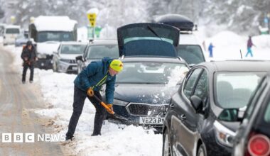 A person in winter clothing shovels snow away from a car on a congested, snow-covered road, with several vehicles stuck in traffic and heavy snowfall visible in a mountainous or rural setting.
