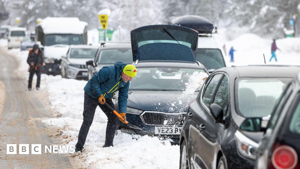 A person in winter clothing shovels snow away from a car on a congested, snow-covered road, with several vehicles stuck in traffic and heavy snowfall visible in a mountainous or rural setting.