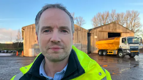 Simon Wells, a man with short grey hair wearing a hi viz jacket, pictured outside several wooden sheds with a large yellow road gritting van