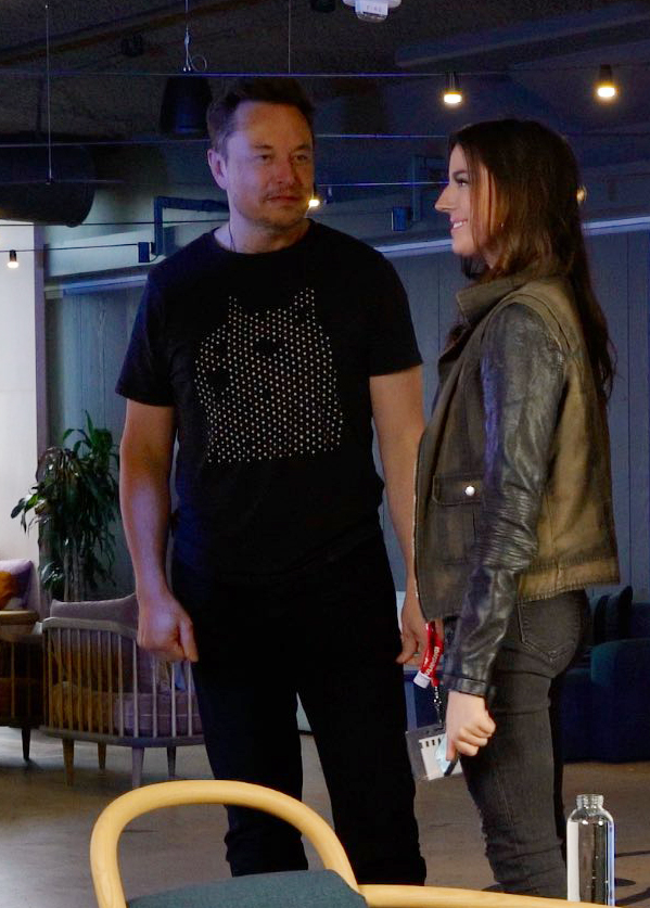 Elon Musk in a black shirt speaks with a woman in a brown and black jacket.
