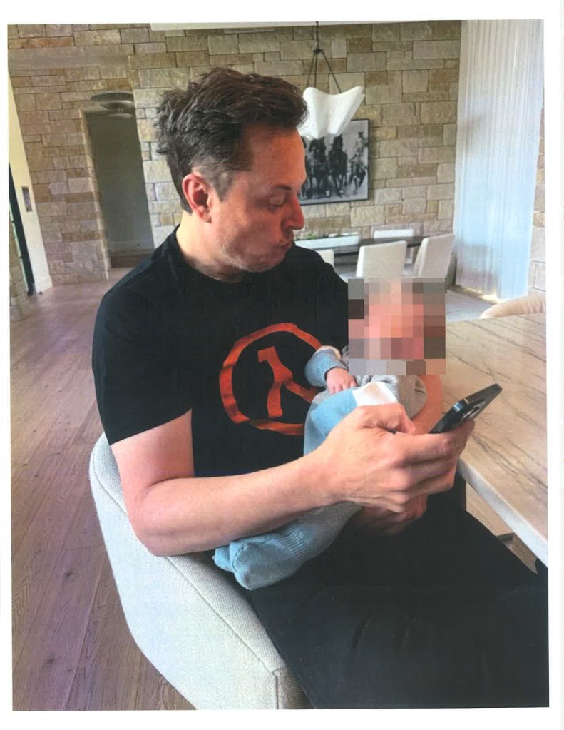 Elon Musk holding a baby whose face is blurred out, while looking at a phone.