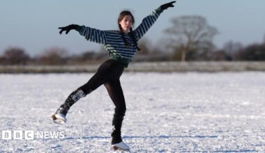 A woman is skating on an icy field. She has long, dark hair in plaits and is wearing a stripey top and trousers. She is a figure skater and her arms are elegantly outstretched as she skates with one foot on the ice and the other raised