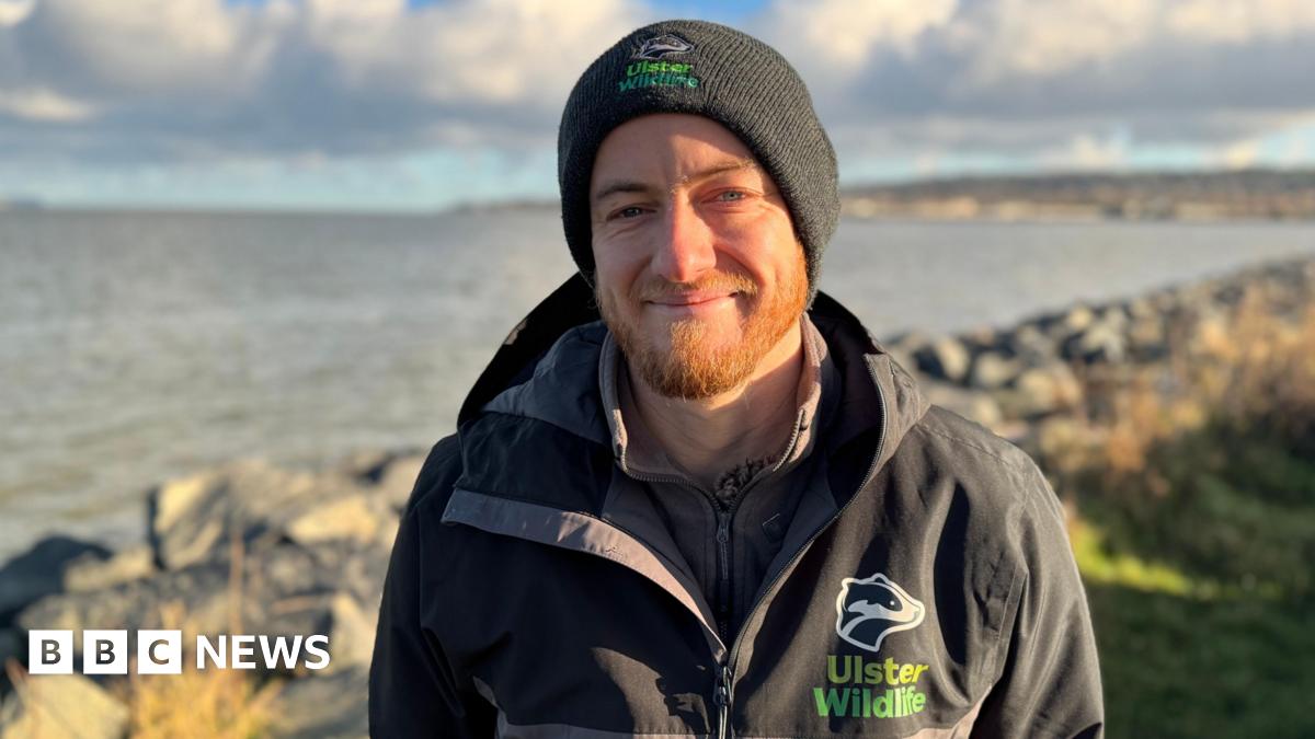 A man standing in front of Belfast Lough. He is wearing a black and grey rain jacket with an Ulster Wildlife logo on it. He is also wearing a black hat.