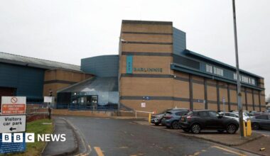 A large prison building, with a car park next to it. The building is sandstone coloured, with a sign saying Barlinnie in large letters on the wall