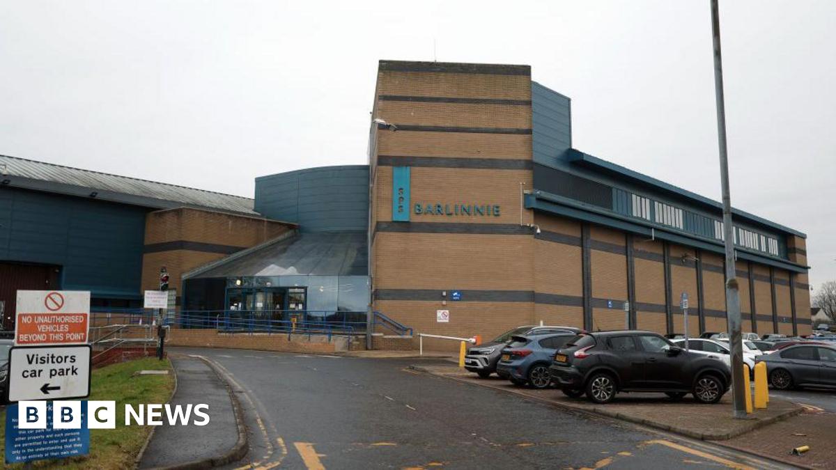 A large prison building, with a car park next to it. The building is sandstone coloured, with a sign saying Barlinnie in large letters on the wall