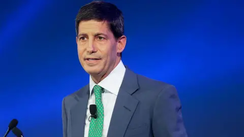 Bloomberg via Getty Images Kevin Warsh, wearing a blue suit and green tie stands in front of a podium with a blue background behind him