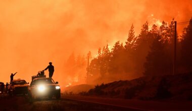Feuerwehrleute stehen auf einem Lastwagen, während im Hintergrund Waldbrände lodern. Der Himmel und die raucherfüllte Luft sind orangefarben von den Flammen erleichtet.