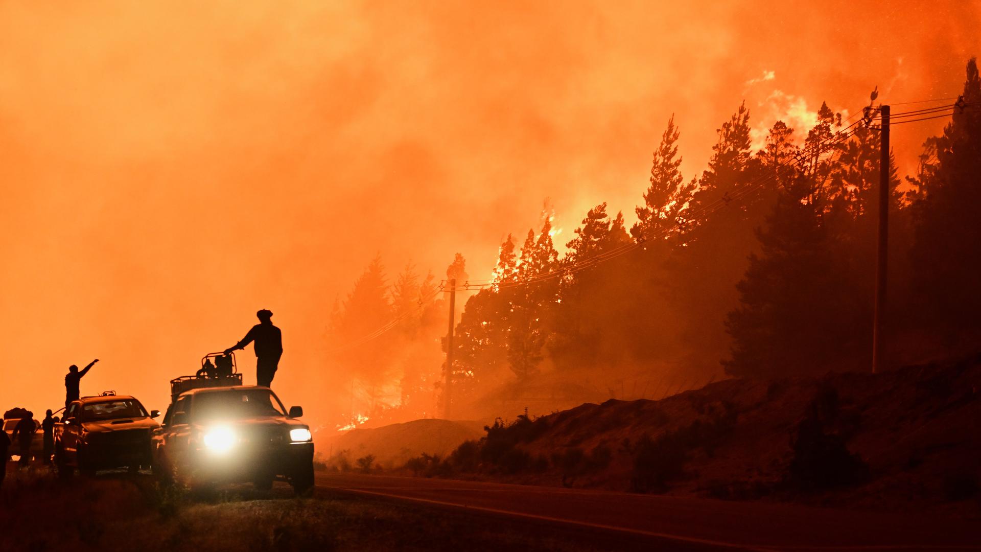 Feuerwehrleute stehen auf einem Lastwagen, während im Hintergrund Waldbrände lodern. Der Himmel und die raucherfüllte Luft sind orangefarben von den Flammen erleichtet. Feuerwehrleute stehen auf einem Lastwagen, während im Hintergrund Waldbrände lodern. Der Himmel und die raucherfüllte Luft sind orangefarben von den Flammen erleichtet.