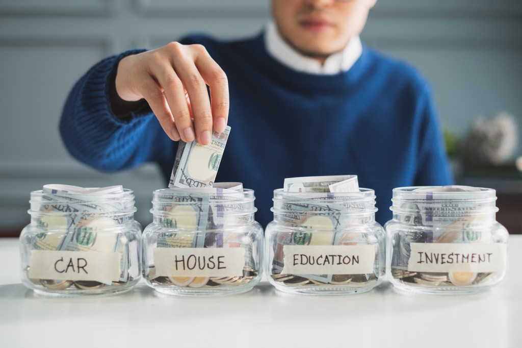 Man putting money into a jar labeled "House," among jars labeled "Car," "Education," and "Investment."