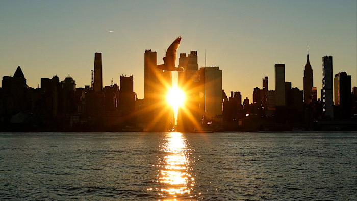 The sun rises behind midtown Manhattan’s skyline with a bird silhouetted in front and its reflection on the Hudson River.