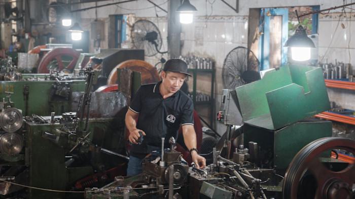 A worker operates a screw manufacturing machine surrounded by industrial equipment at a factory in Binh Duong Province, Vietnam.