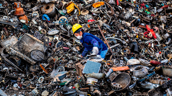 A worker wearing a yellow hard hat and face mask sorts through a large pile of mixed scrap metal at a recycling plant.