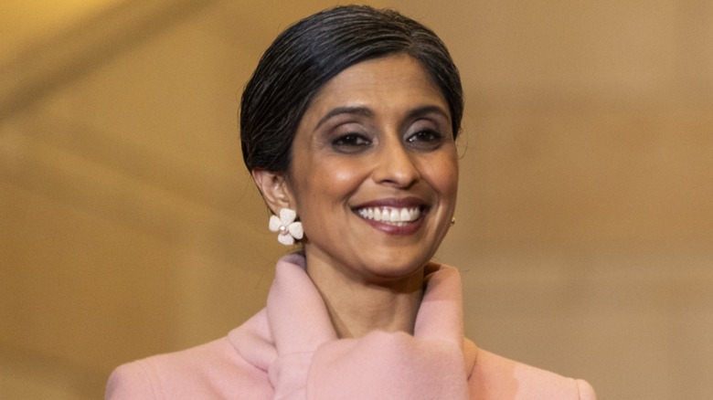 Wearing a pink coat with knotted detail, Usha Vance smiles while gazing downward.